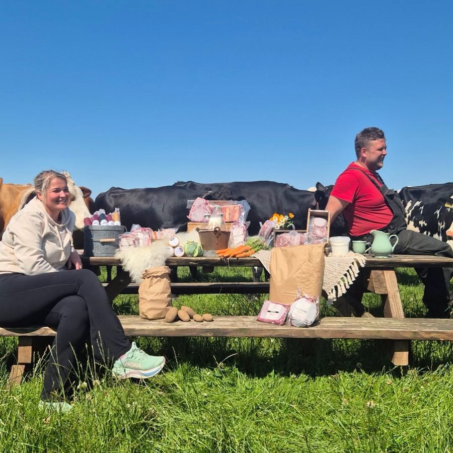 Familien bak Hole Gardsutsalg ved piknikbordet ute på jordet, med varer på bordet og kyr bak.
