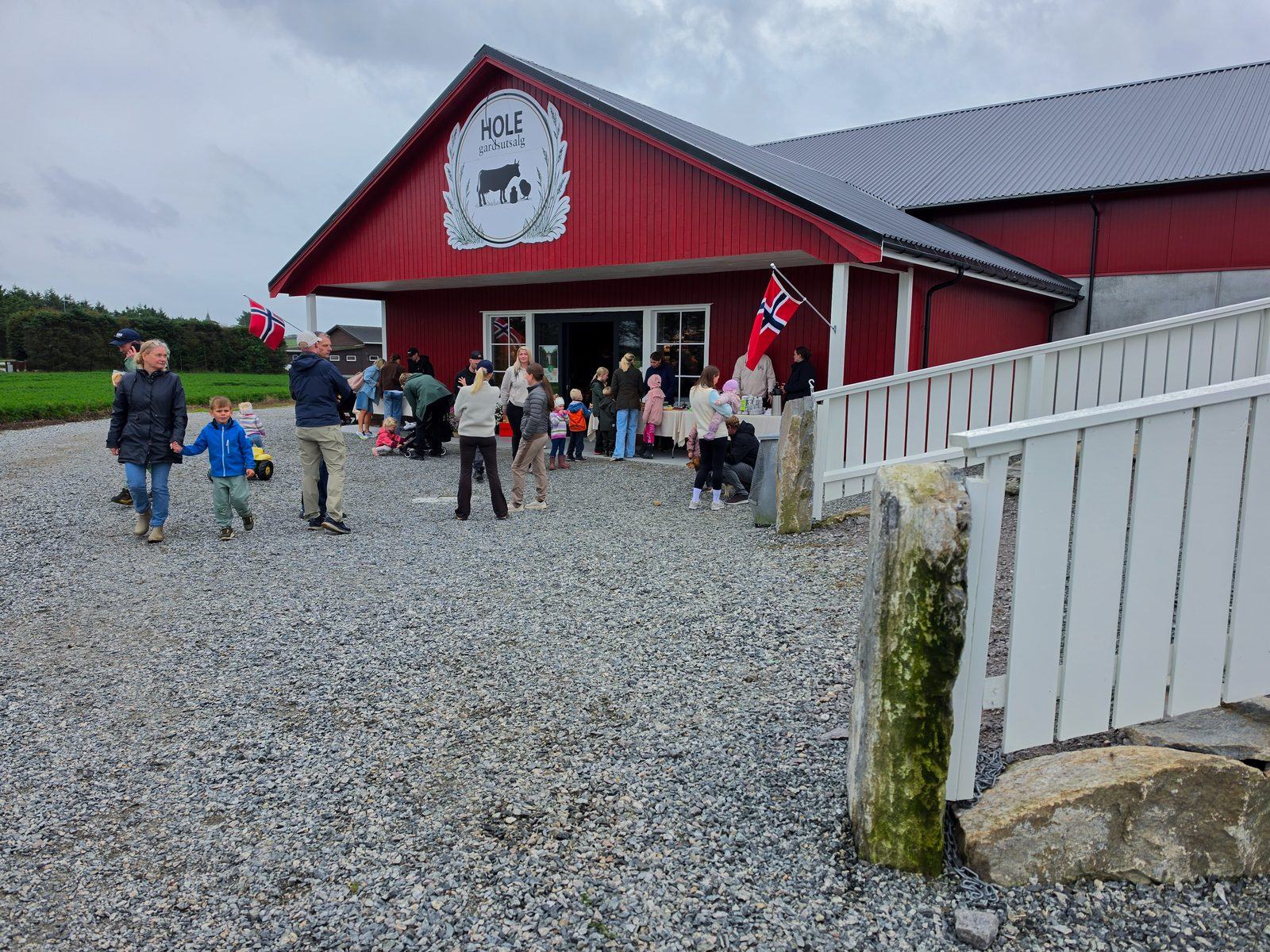 Folk og familier utenfor gårdsutsalget til Hole Gardsutsalg på en travel dag – rødt bygg med HOLE-emblemet og norsk flagg.